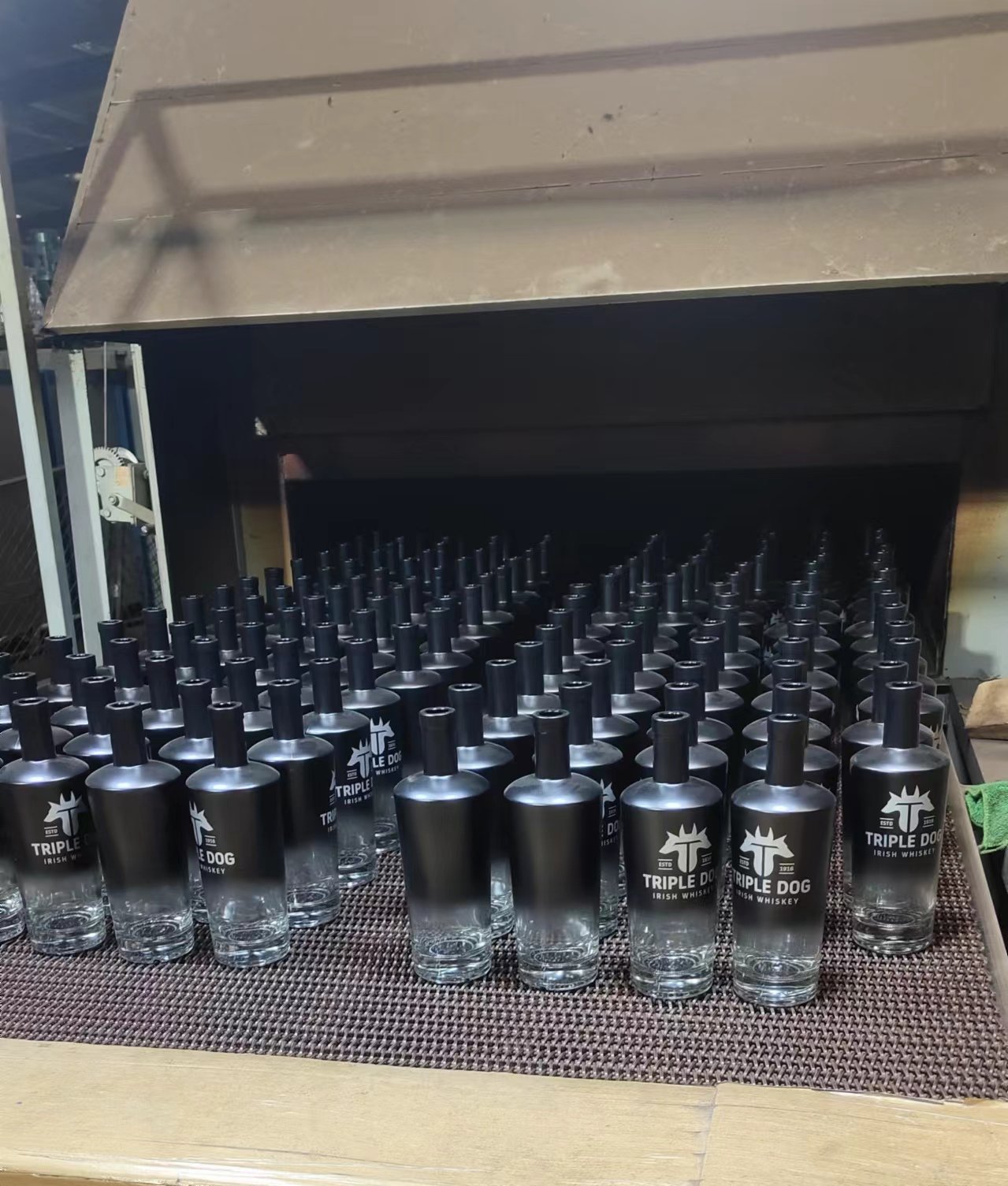 Rows of matte-sprayed, screen-printed spirit bottles entering a curing oven on the conveyor.